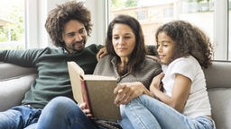 Vater mit dunkler Hautfarbe, seine weiße Frau und ihre gemeinsame Tochter sitzen auf dem Sofa und lesen zusammen in einem Buch.