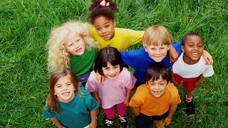 Kinder mit verschiedenen Hautfarben stehen auf einer Wiese und gucken nach oben in die Kamera.