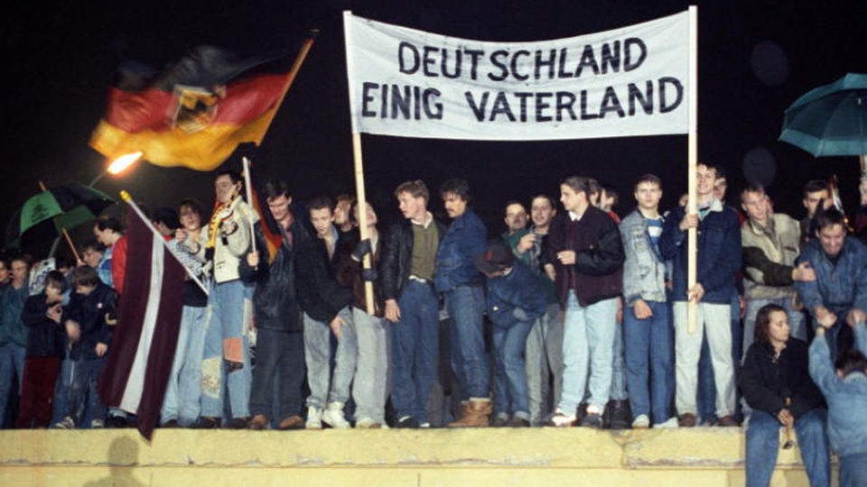 Berliner stehen mit der Deutschlandfahne und einem Transparent &#8218;Deutschland Einig Vaterland&#8217; auf der Berliner Mauer.