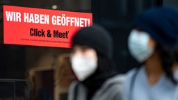 Zwei Frauen mit Mund-Nasen-Schutz vor Schaufenster mit Schild 'Wir haben geöffnet - click & meet'.