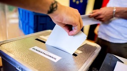 Hand steckt Wahlzettel in Urne mit der Aufschrift 'Bundestagswahl'.