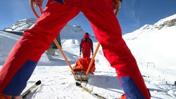 Beine eines Bergretters in roter Kleidung vor einer Trage, die im Schnee geschoben wird.