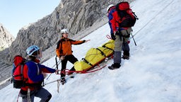 Bergwachtanwärter mit großen Rucksäcken sichern Verletzten in einer Trage auf verschneitem Berg.