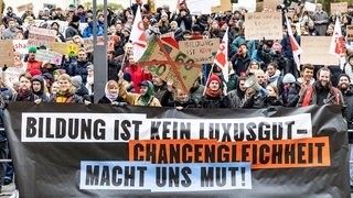 Demonstranten halten Pappschilder hoch, im Vordergrund Transparent mit der Aufschrift "Bildung ist kein Luxusgut, Chancengleichheit macht uns Mut".