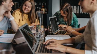 Mädchen sitzen mit mehreren Laptops an Tisch und lernen gemeinsam.