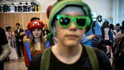 Jugendlicher Cosplayer mit grüner Brille, im HG Cosplayerin, die als Super-Mario verkleidet ist.