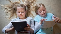 Zwei Mädchen liegen nebeneinander auf dem Rücken, beide schauen auf ein Tablet.