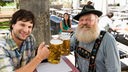Zwei Männer prosten sich mit Bier zu, einer trägt bayrische Tracht und Bart.