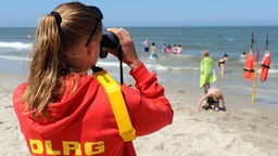 Eine junge Rettungsschwimmerin der DLRG überwacht mit einem Fernglas einen Strandabschnitt. 