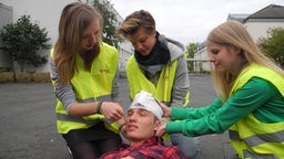Drei Schulsanitäter in gelben Westen kümmern sich in einer Übung um einen Schüler mit Kopfverband.