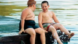 Zwei übergewichtige Jungen sitzen in Badehosen auf einem Steg.
