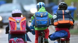Drei Kinder (von hinten) fahren mit Fahrrädern nebeneinander her, alle drei tragen Schulranzen und Fahrradhelme.