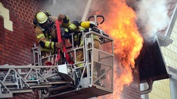Zwei Feuerwehrleute in Rettungskorb vor brennendem Haus.