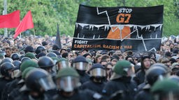Demonstranten tragen ein Plakat auf dem &#8222;Stop G7&#8220; steht, im Vordergrund sind Polizisten mit Helmen.