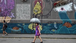 Frau mit Regenschirm geht an einer Wand voller Graffiti vorbei.