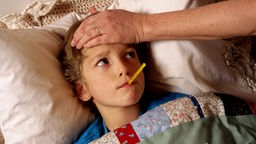 Ein Kind liegt krank im Bett und hat ein Thermometer im Mund.