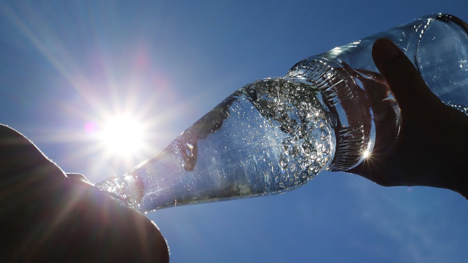 Mensch trinkt aus Wasserflasche, im Hintergrund blauer Himmel und Sonne.