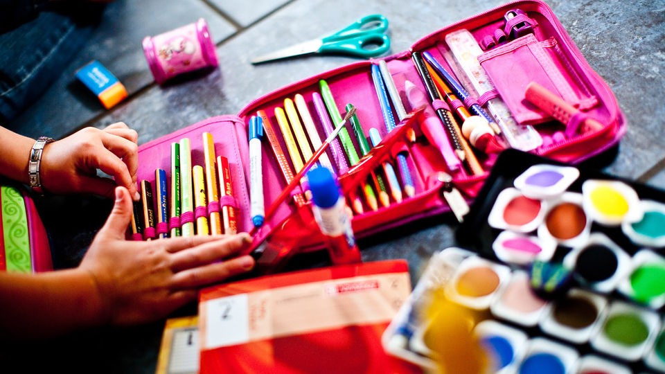 Geöffnetes Federmäppchen mit Stiften, Wasserfarbkasten und weitere Schulmaterialien liegen auf einem Tisch.