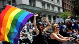 Zwei Frauen sitzen auf einem Motorrad, die hintere schwenkt eine Fahne in Regenbogenfarben.