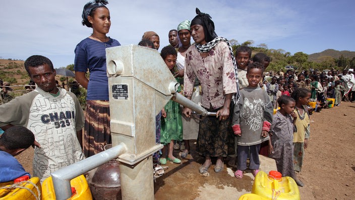 Eine Frau pumpt Wasser aus einem Brunnen, dahinter warten Menschen in der Schlange.