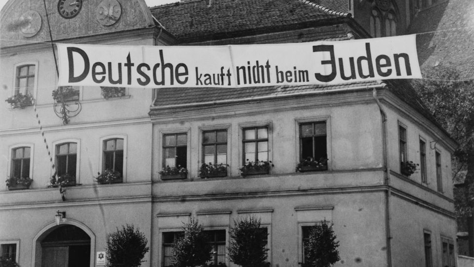 Banner mit der Aufschrift 'Deutsche kauft nicht beim Juden' hängt an Gebäude.
