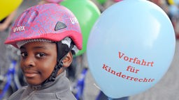 Junge mit rosanem Fahrradhelm neben hellblauem Luftballon mit der Aufschrift 'Vorfahrt für Kinderrechte'.