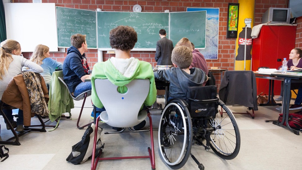 In einem Klassenraum sitzt ein Schüler im Rollstuhl neben einem Schüler auf einem Stuhl, beide sieht man von hinten.