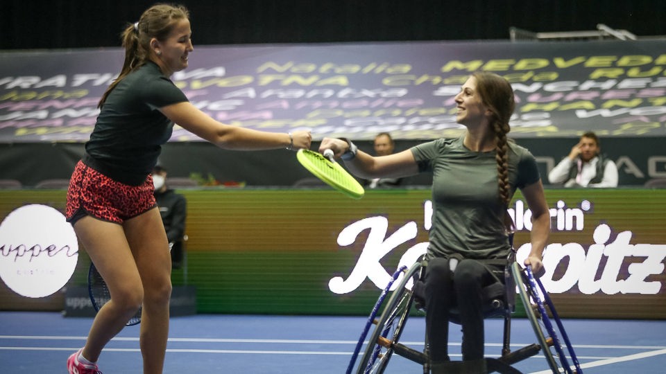 Zwei Tennisspielerinnen begrüßen sich auf dem Tennisplatz. Eine Spielerin sitzt im Rollstuhl.