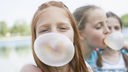 Zwei Mädchen stehen hintereinander, beide haben eine dicke Kaugummiblase vor dem Mund.