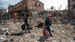 Zwei Frauen mit großen Taschen gehen vor zerstörten Gebäuden.
