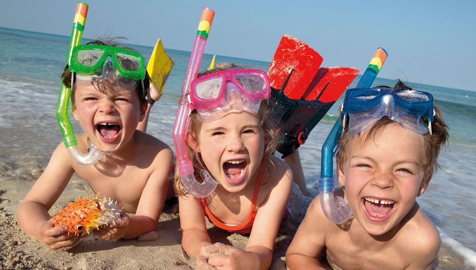 Drei Kinder mit Schnorcheln liegen an Strand und lachen.
