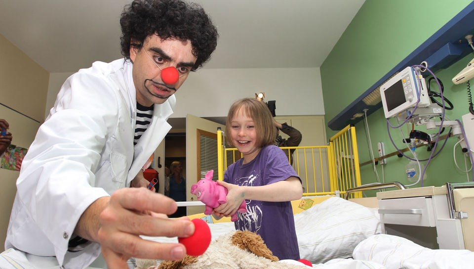 Ein Clown mit roter Nase spielt mit einem Mädchen in Krankenhauszimmer.