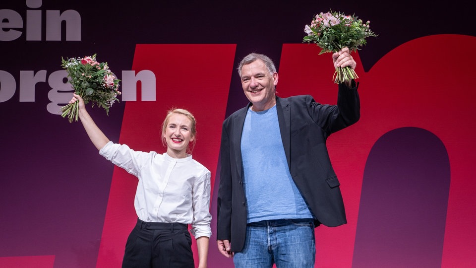 Die Parteivorsitzenden Ines Schwerndtner und Jan van Aken winke auf der Bühne mit Blumensträußen in der Hand.