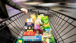 Hand an einem Einkaufswagen mit verschiedenen Lebensmitteln.