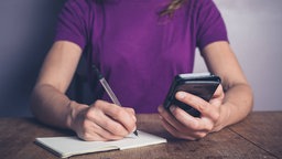 Eine junge Frau sitzt am Tisch und schreibt in ein Notizbuch.