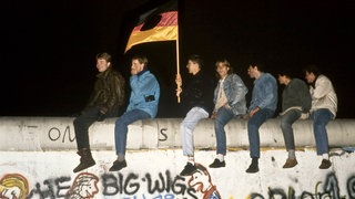 Sieben Männer sitzen rittlings auf der Berliner Mauer und halten eine Fahne, aus der das Emblem der DDR herausgeschnitten wurde.