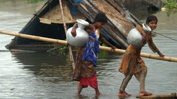 Zwei Mädchen tragen große Gefäße und balancieren über ein Holzbrett, das über einem überschwemmten Gebiet liegt.