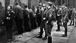 Nationalsozialisten in Uniformen der SA drängen Männer an eine Mauer.