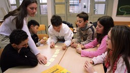Kinder mit Migrationshintergrund sitzen an Tisch, daneben Lehrerin.