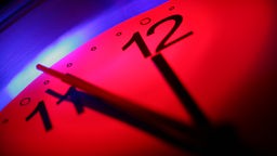 Ausschnitt Uhr mit rotem Ziffernblatt, Zeiger stehen auf fünf vor zwölf.