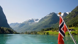 Fahne weht an Heck von Boot, dass durch norwegischen Fjord fährt.