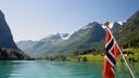 Fahne weht an Heck von Boot, dass durch norwegischen Fjord fährt.