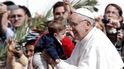 Papst Franziskus hebt ein kleines Kind hoch.