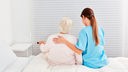 Eine Pflegerin (von hinten) sitzt mit einer alten Dame auf dem Bett und hat einen Arm auf ihren Rücken gelegt.