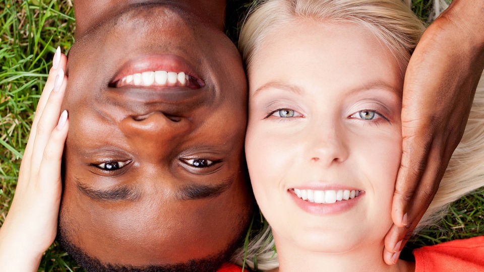 Der Kopf eines dunkelhäutigen Mannes liegt neben dem Kopf einer Frau mit heller Hautfarbe. Beide lächeln in die Kamera.