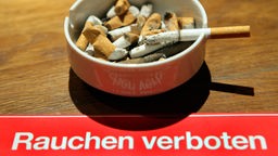 Gefüllter Aschenbecher, darunter ein Schild mit der Aufschrift "Rauchen verboten".