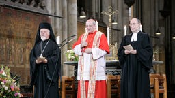 Ein griechisch-orthodoxer Bischof, ein katholischer Erzbischof und ein evangelischer Theologe stehen in einer Kirche nebeneinander.