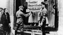 Zwei SA-Mitglieder hängen ein Schild mit der Aufschrift &#8222;Deutsche! Wehrt Euch! Kauft nicht bei Juden&#8220; an ein Schaufenster