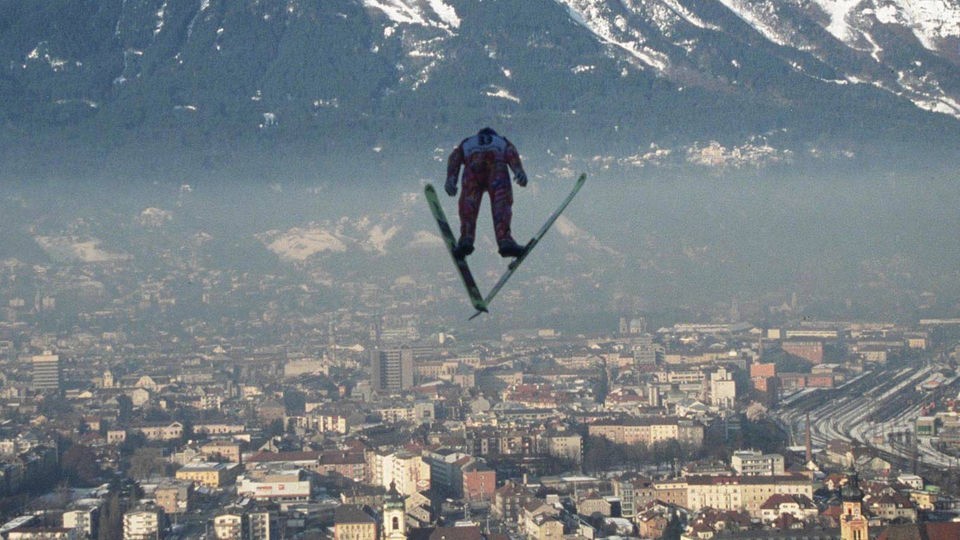 Skispringer im Flug hält Skier in V-Form, im Tal sieht man die Stadt Insbruck.
