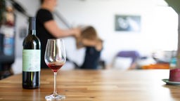 Im Vordergrund stehen ein Weinglas und eine Weinflasche, im Hintergrund hält ein Erwachsener ein Kind fest.